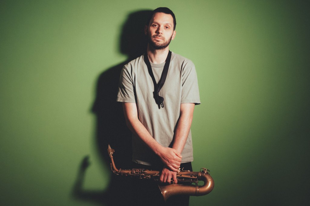 A man stands in front of a green background holding a saxaphone.