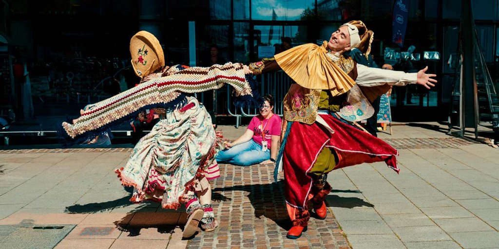 Two dancers hold hands and are swinging each other around. One has a straw bonnet on and the other a jester's hat. They are outside and it's a sunny day.